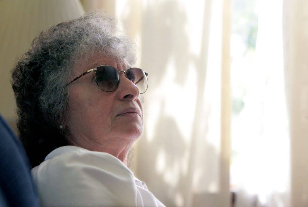 An older woman with curly, shoulder-length gray hair and sunglasses looks off-camera.