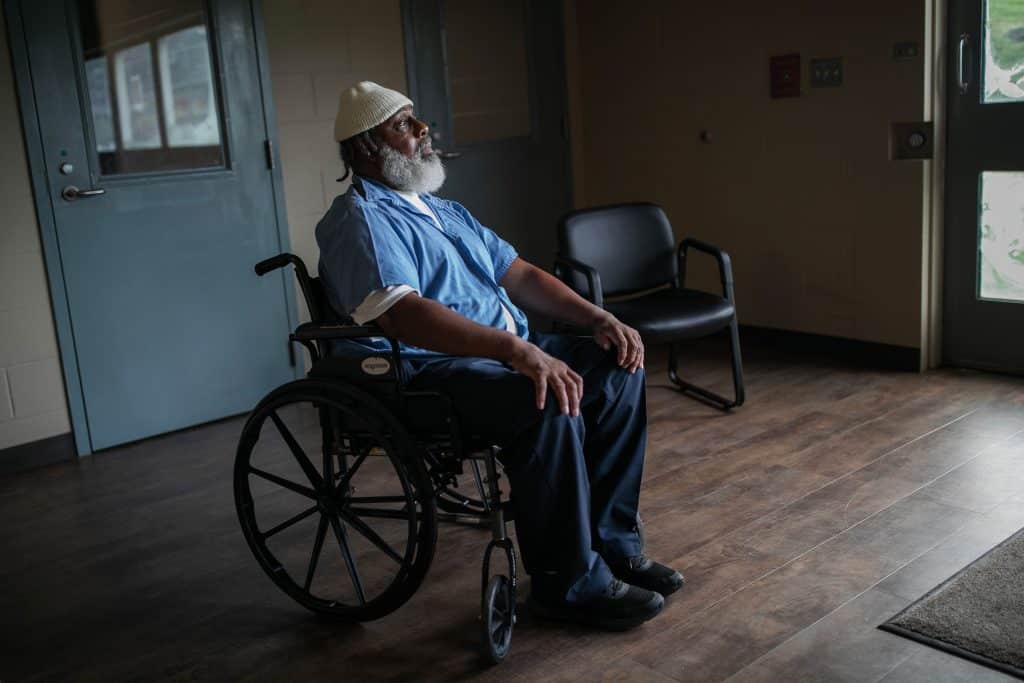 An older Black man — a wheelchair user with a gray beard, white knit hat and blue shirt — looks off camera.