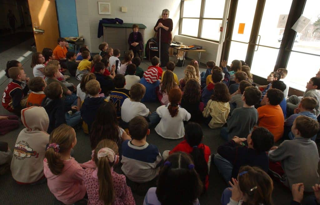 An older woman with curly, shoulder-length gray hair, sunglasses and a cane speaks in front of a room of more than 50 elementary students sitting on the floor in front of her.