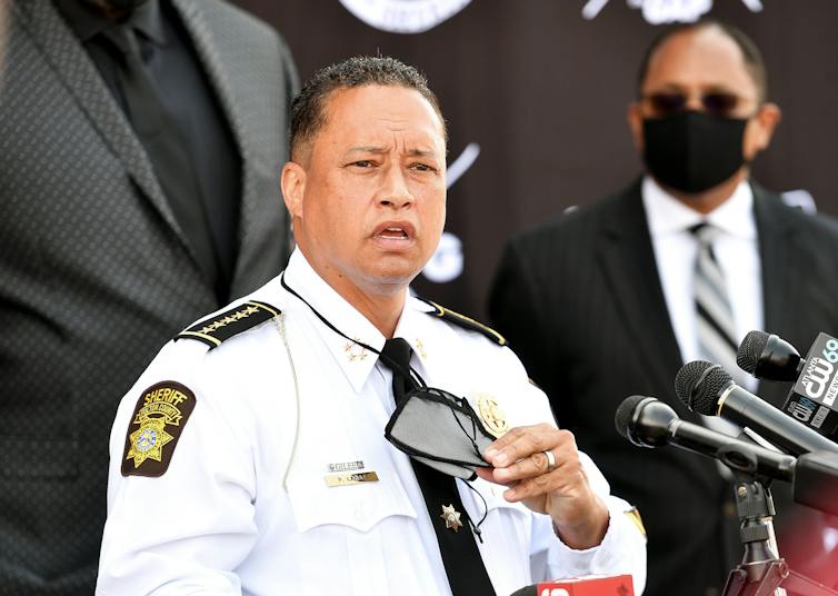 A middle aged man dressed in a white shirt adorned withlaw enforcement patches is speaking to a crowd.