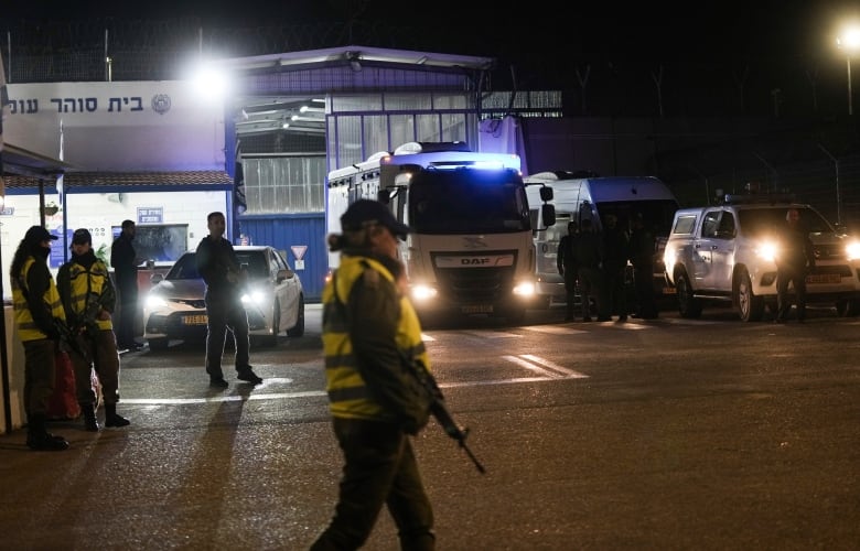 A guard wearing a neon yellow vest and holding an long gun stand in front of a bus at a checkpoint at night. 