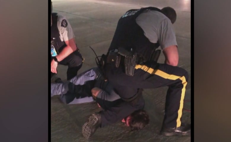 Two uniformed RCMP officers hold down a man lying on a sidewalk, with one of the officers' knees on the man's neck.