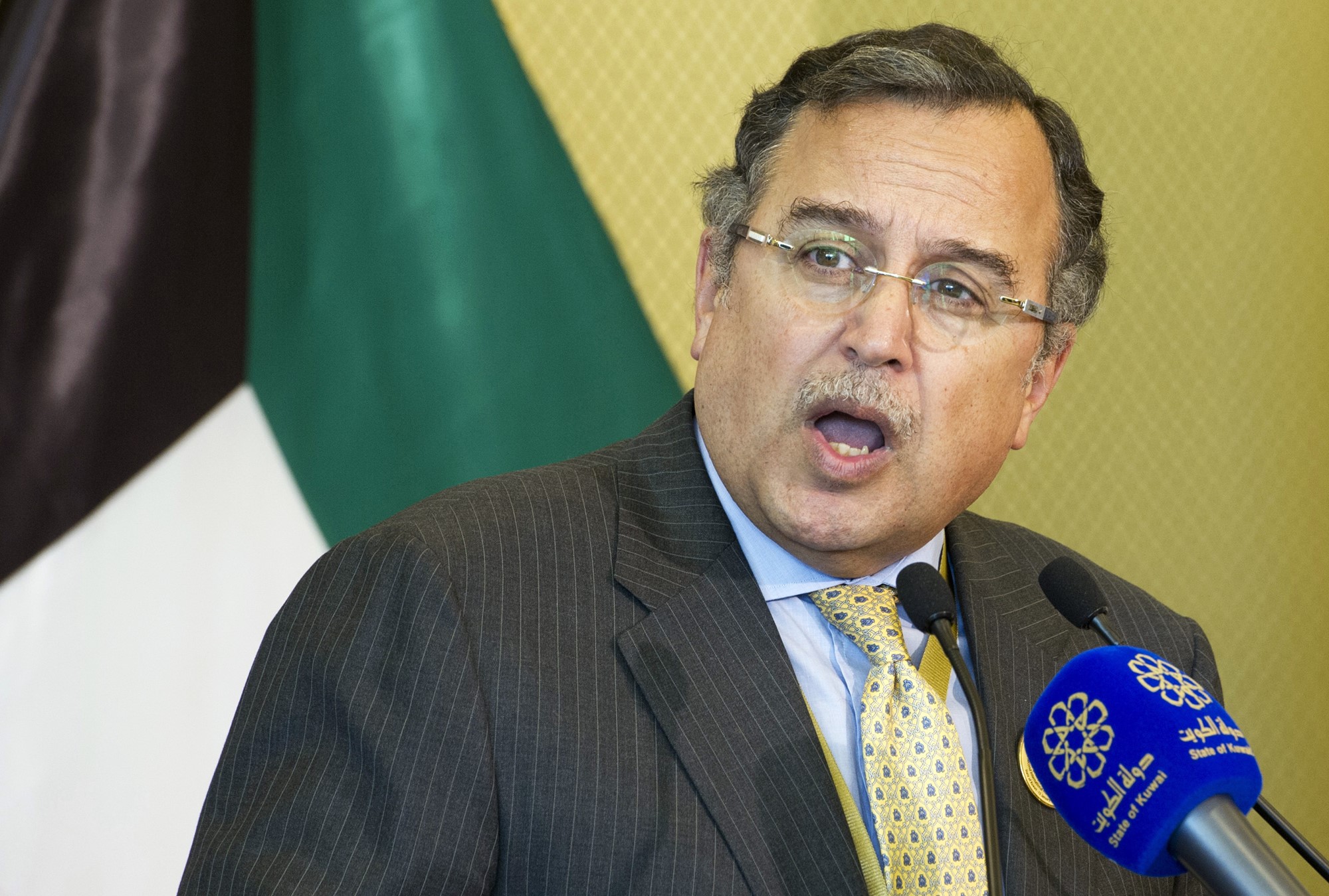 Man in brown suit with yellow tie and glasses talks into a microphone