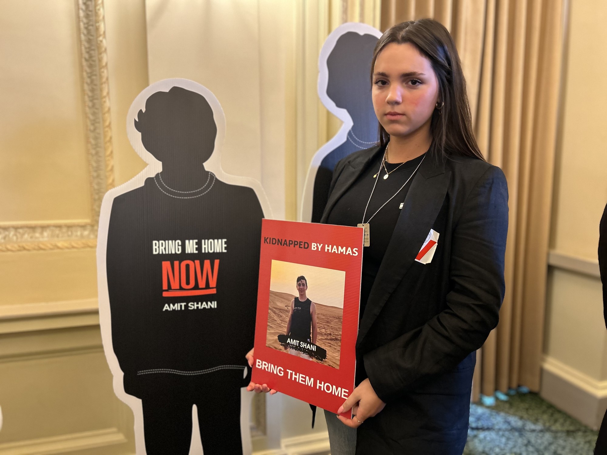 A girl in a black suit holding a read sign with an image of a boy on it with two cardboard cutouts of silhouette of children behind her