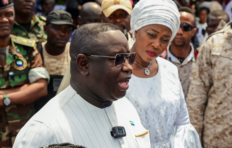 A man with sunglasses speaks in the foreground, while a woman in a headwrap listens behind him. Surrounding them are people in military uniform.