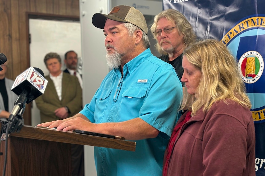 A man wearing a button-up t-shirt and a cap speaks to the press, a man and a woman stand behind him