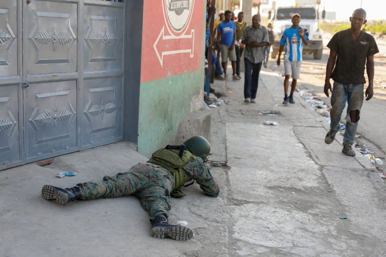 Pedestrians walk past a soldier in uniform lying on the ground.