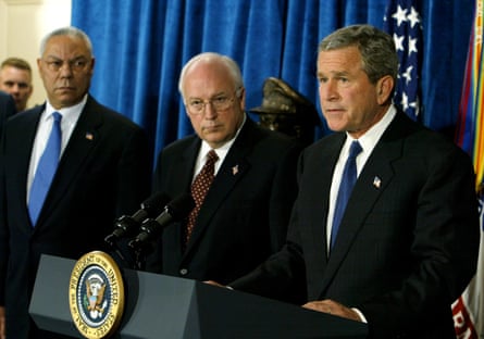 Standing in a row, three middle-aged men in suits, one Black and two white. The man on the furthest right speaks into a microphone.