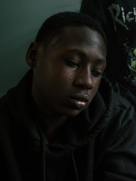 A young man Black, eyes downcast, with short black hair and a black sweatshirt, appearing to sit against a wall inside somewhere.