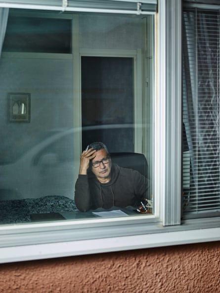 Middle-aged Arab man with short gray hair in sweater is seen through the plate-glass window of a house, holding a pen and looking through the window.