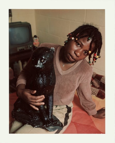 A woman with braided and beaded hair posing with a large bear ornament on the bed in her cell