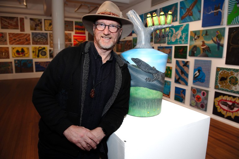 Sean Miller. He is standing in front of a ceramic art work. He's wearing a black short and coat as well as a hat.