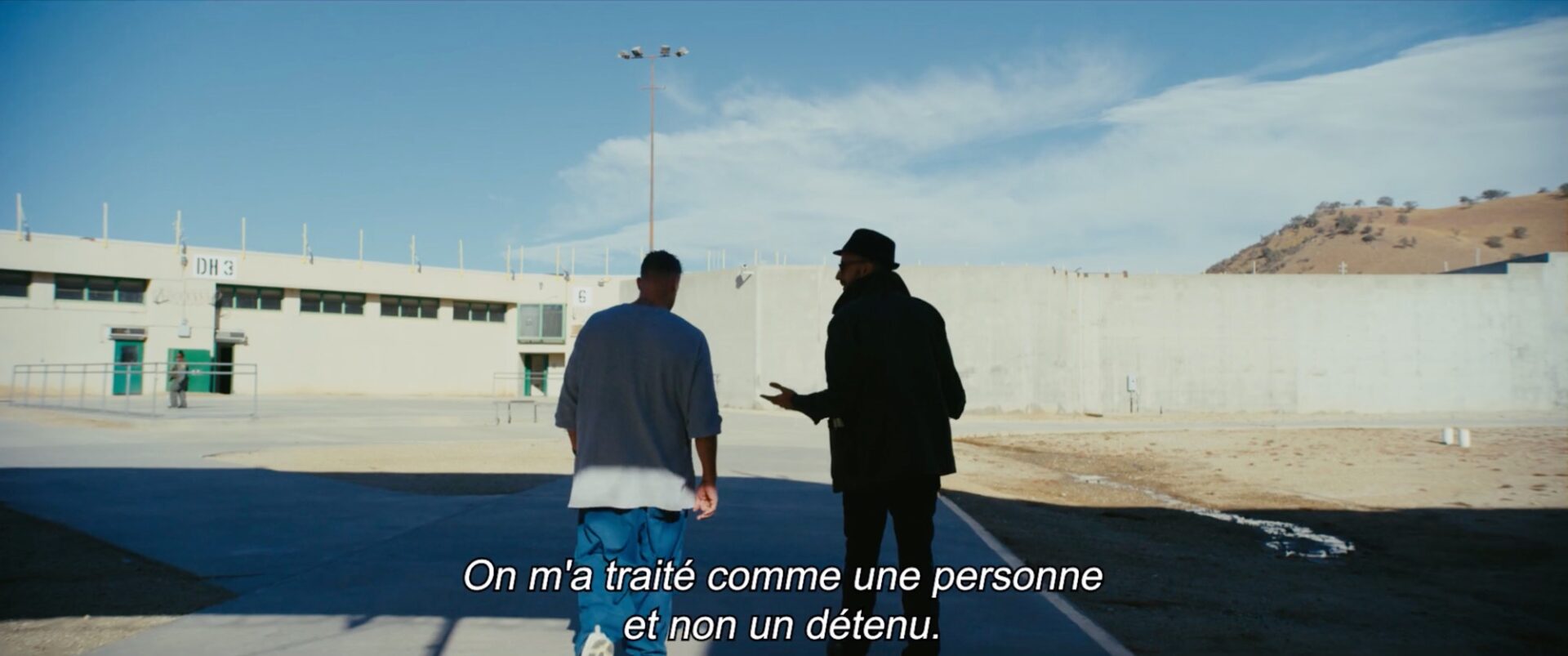 a still from a documentary in a prison in California showing artist JR walking with an inmate through the prison yard, with a French substitle translated to 