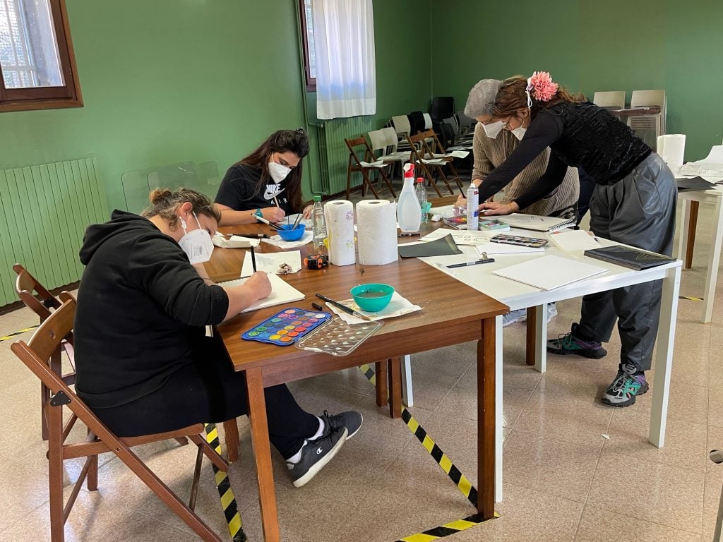 a group of women wearing covid masks sit at a table doing crafts and drawing in a room with green painted walls