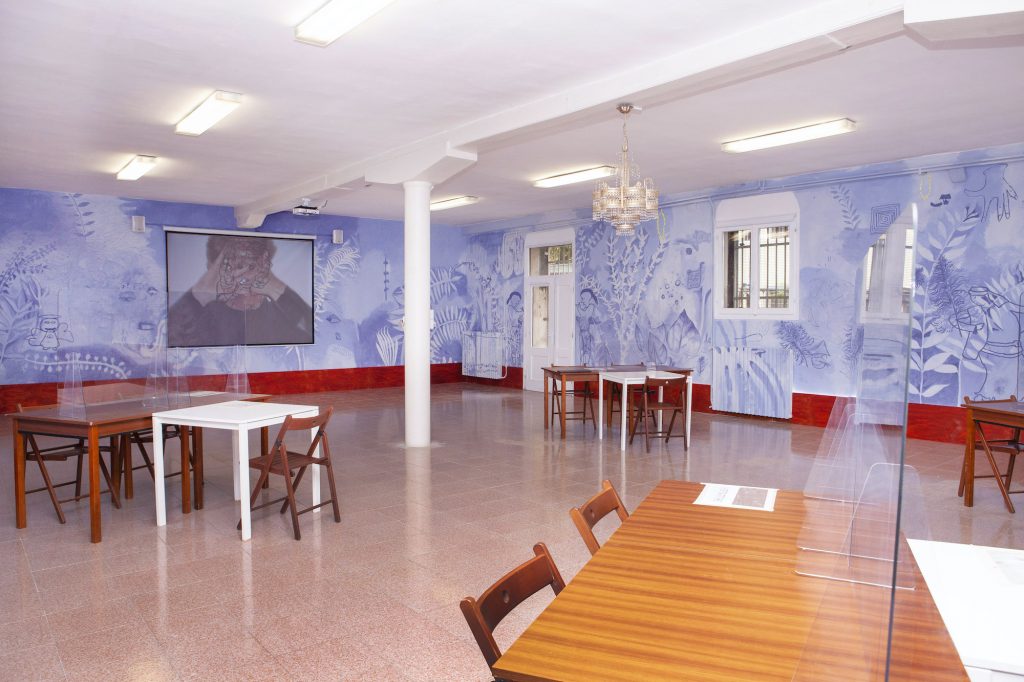 a large room with tables has a light blue and purple mural on the walls and a screen showing a video installed on one wall