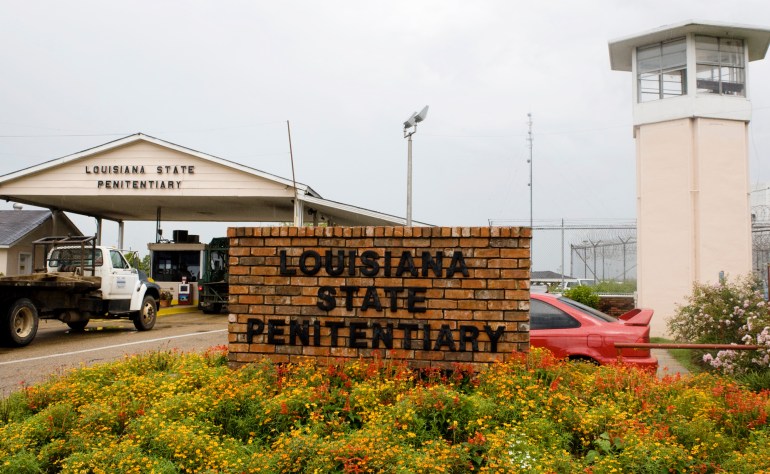 A view of the entrance of the Louisiana State Penitentiary in Angola. A watch tower rises on one side, next to a covered entrance way with a security checkpoint that vehicles pass through. In the front sits a brick wall with the words: Louisiana State Penitentiary.