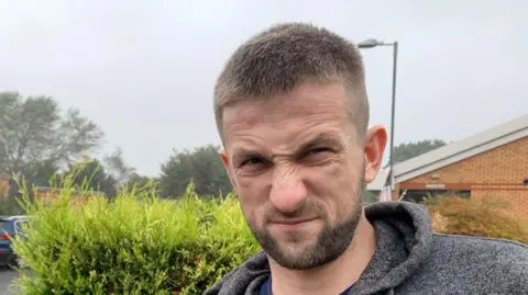 Mick Lunney/BBC Jake Crowder, who has short hair and a beard, looks into the camera. He is wearing a grey hooded top and standing in front of a hedge