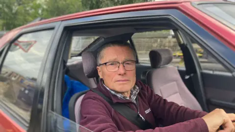 Steve Jones/BBC Andrew sits in a car dressed in a burgundy fleece waiting for his son