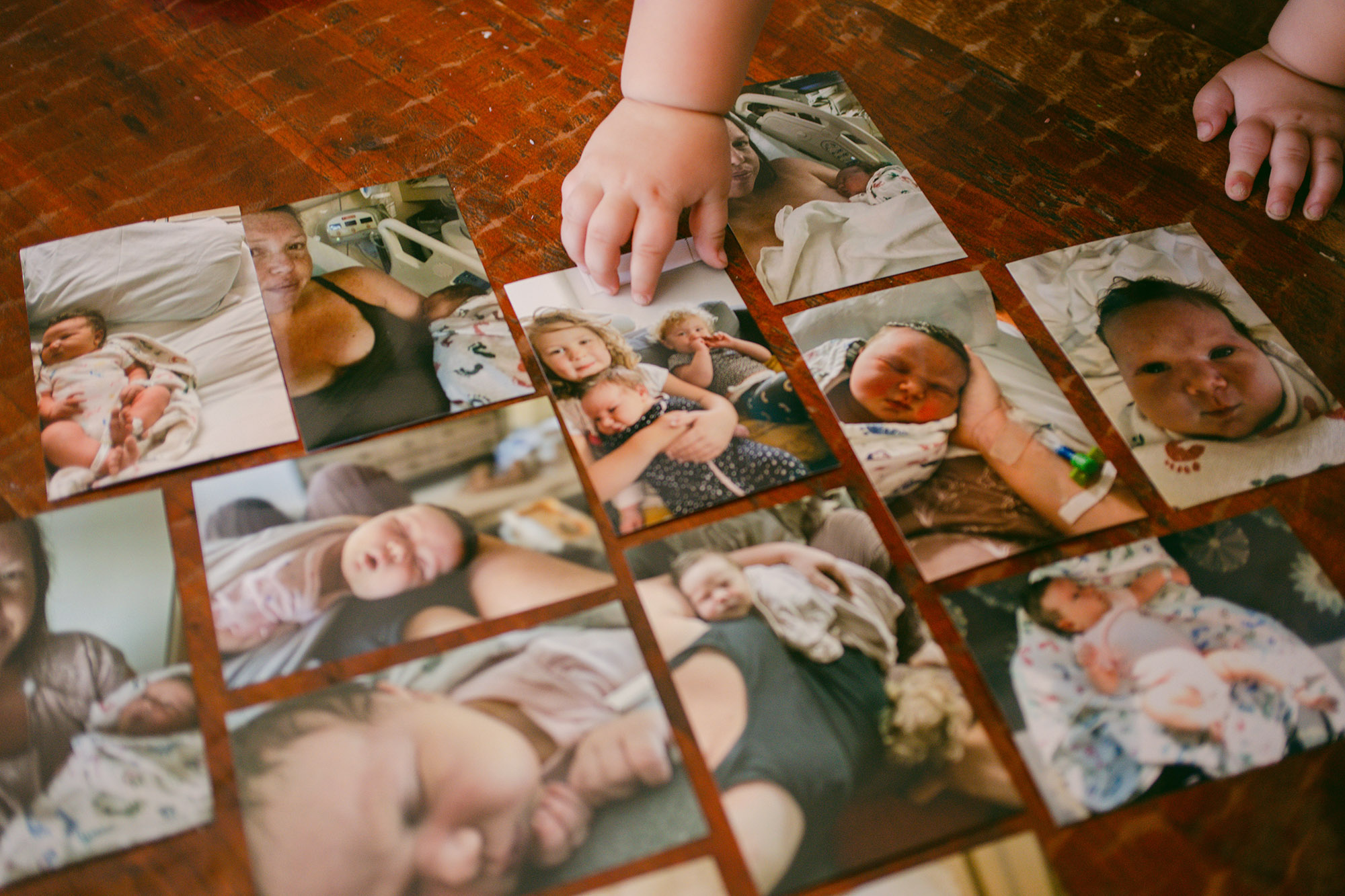 Twelve photos are arranged on a wooden floor showing a woman with her newborn and two of her other children. A toddler's hand is touching one of the photos.