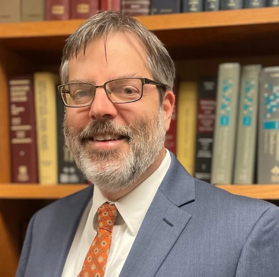 Cayuhoga juvenile defenders: Middle-aged man with salt and pepper hair and full beard with glasses in gray suit, white shirt and red tie, in front of wood book shelves