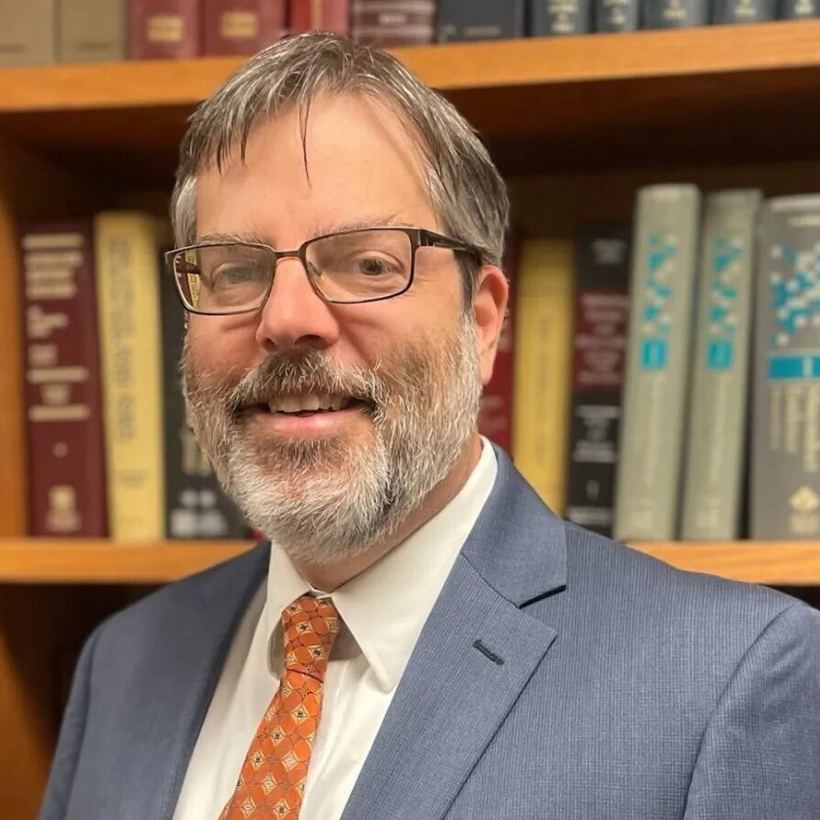 Cayuhoga juvenile defenders: Middle-aged man with salt and pepper hair and full beard with glasses in gray suit, white shirt and red tie, in front of wood book shelves