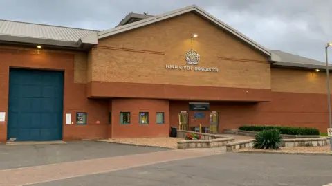 BBC/Mick Lunney Brick-built HMP Doncaster with small all around entrance path and larger blue door for exits to the left of the image