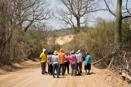 A group of people seen from behind stand on a dirt road looking at a digger and a dumper truck on cleared land in the distance 