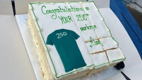 A cake saying 'Congratulations on your 250th parkrun Alan' with a green shirt on it reading '250'