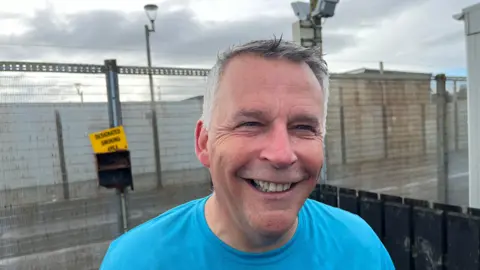 BBC Gary Milling, governor of Magilligan Prison. He is smiling, has silver hair and wearing a blue t-shirt. Outside fencing of the prison can be seen in the background.