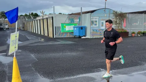 Prison officer Colin Rice running alongside part of the prison. He has dark hair and wearing a black t-shirt and black shorts.