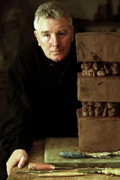 Getty Images Jimmy Boyle, wearing black clothing, standing next to a three-level clay sculpture. It is placed on a table, with tools lying nearby.