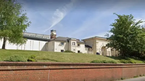 Google A screenshot from Google street view of the outside of the prison. It's a white building behind a grassy hill and a brick wall.