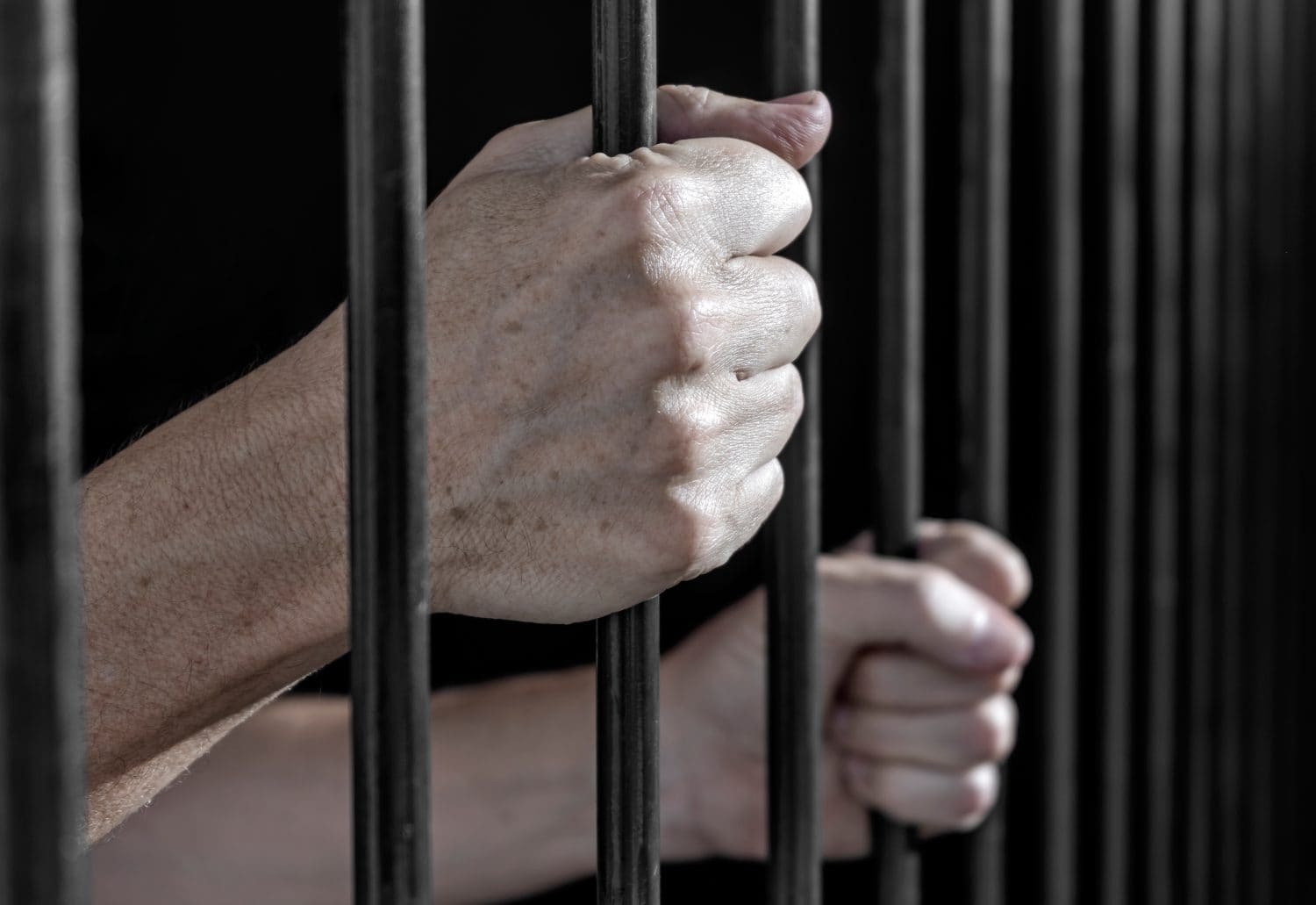 Prisoner's hands gripping the bars of a cell in jail or prison serving sentence.