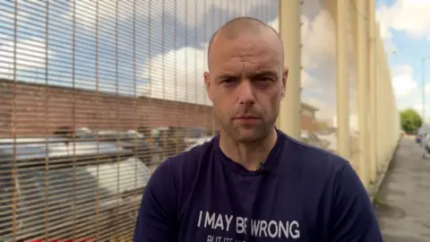 Ben Moore/BBC Jamie is wearing a black t-shirt and is standing next to a tall fence. He has a shaved head and stubble. 