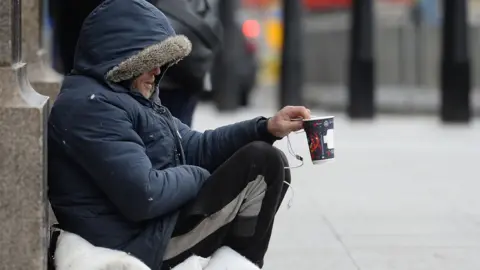 Nicholas.T.Ansell/PA Wire A man wearing a blue parka jacket and black jogging bottoms sits on a duvet on the street while holding out an empty coffee cup to passers by