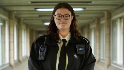 Ministry of Justice Hollie Peabody at work. They wear a black prison officer's uniform including a black tie and jacket over a white shirt. They have a pager and walkie-talkie attached to their jacket and wear dark-rimmed rectangle glasses. Hollie is pictured in a brutalist corridor with windows on either side and looks straight at the camera. 