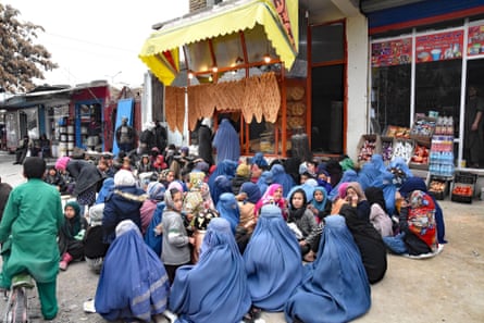 A crowd of women in burqas, some with children, sit on the ground outside a bakery