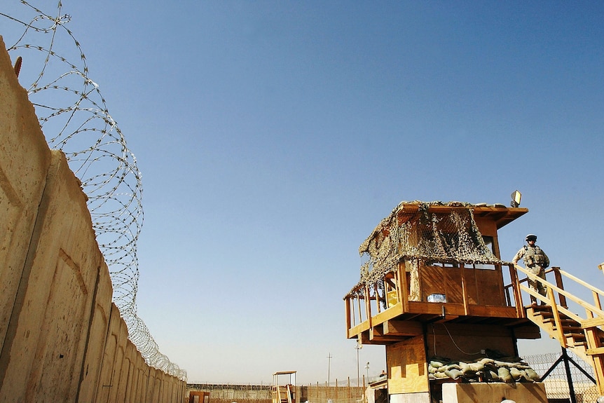 A barbed wire fence of a prison next to a watch tower where a soldier stands guard