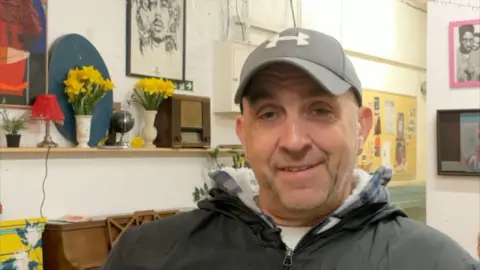 Mr Devonport smiles at the camera. He is wearing an Under Armour cap and anorak. Behind him, several pieces of art can be seen on the wall as well as a shelf which carries two vases with daffodils, a small lamp and a small desk globe.