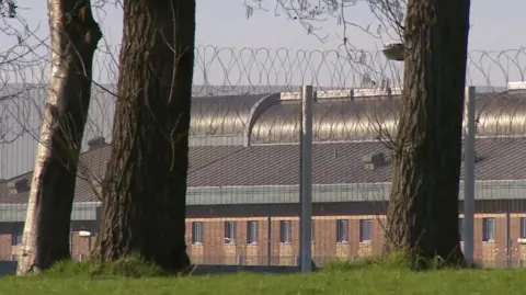 BBC The prison surrounded by a security high fence and barbed wire and two trees in the foreground