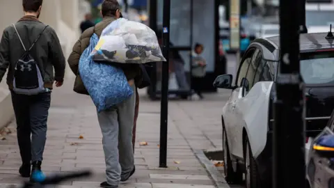 PA Media Two people walk down a pavement, one carrying a large bag of belongings over their shoulder, with a parked car and bus stop in the background.