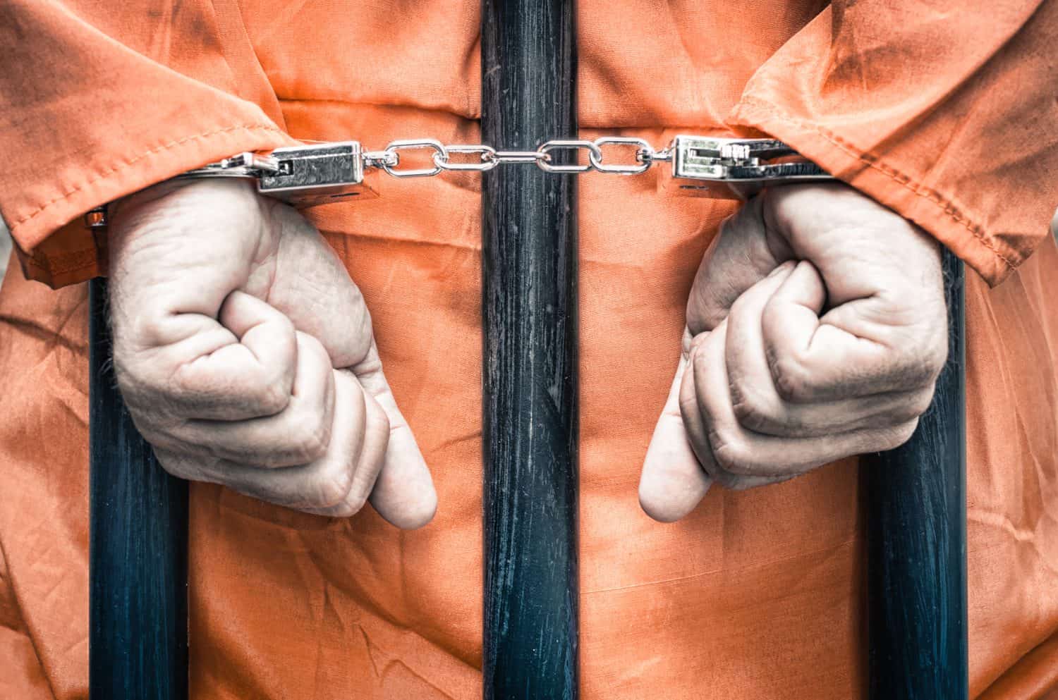 Handcuffed hands of a prisoner behind the bars of a prison with orange clothes - Crispy desaturated dramatic filtered look