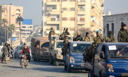 Anti-government fighters sit and stand on vehicles driving through a city street