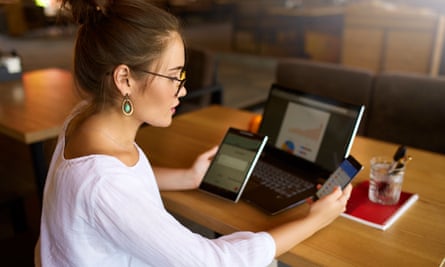 A person looks at smartphone while holding a tablet and sitting in front of a laptop with bar and pie charts on the screen