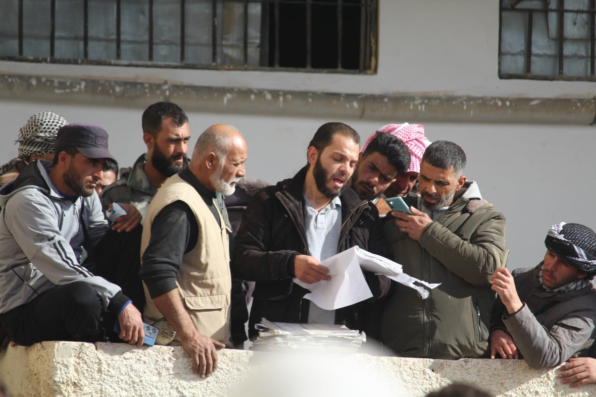 A man surrounded by others reads from a list of names.