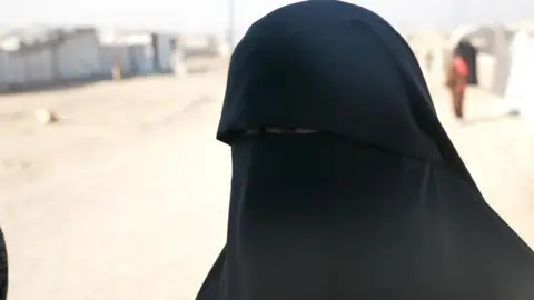 A woman with her head covered in black cloth, her eyes barely visible, outdoors in bright sun in the al-Hol camp in Syria