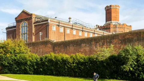 Getty Images Red brick building, behind a tall red brick wall with green hedges in front of it. This is Reading Jail.