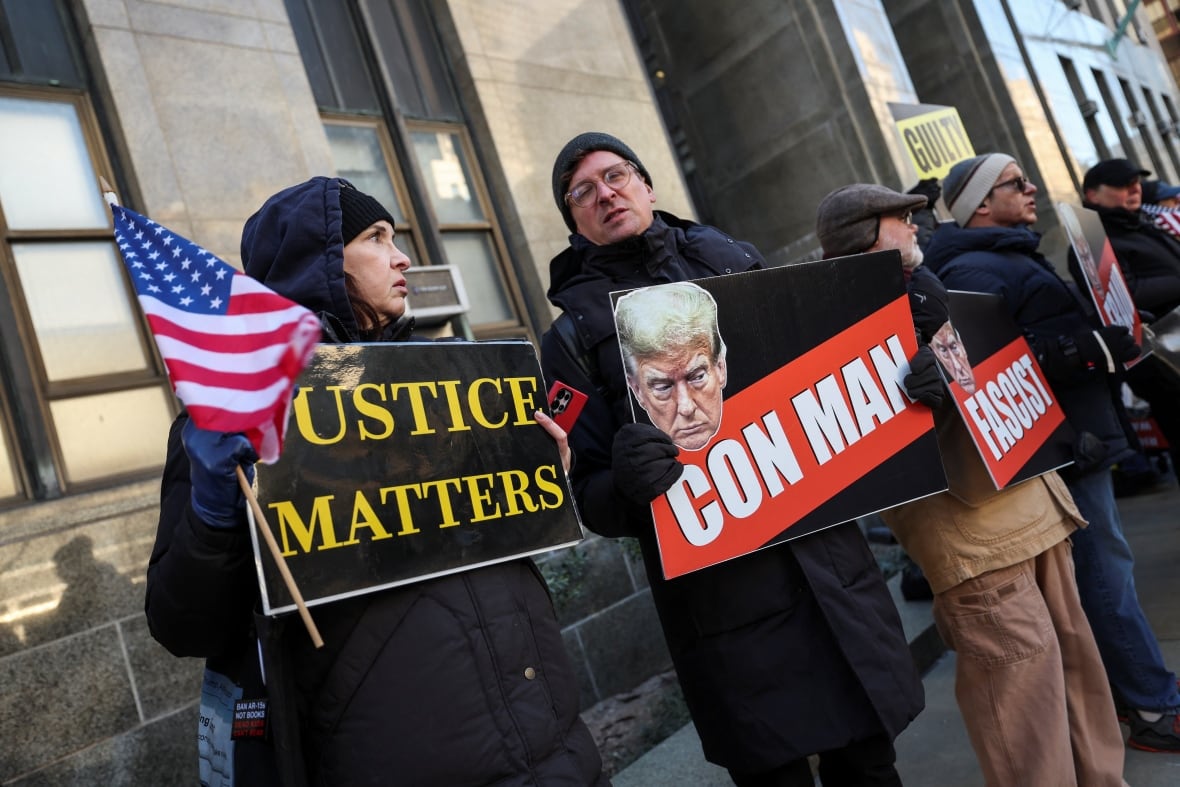 A woman and man in winter clothing hold up signs at an outdoor protest. One sign says 'justice matters,' the other says 'con man.'