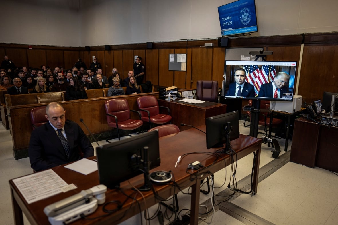 An attorney is seen sitting at a table in a courtroom while two other men appear via video.