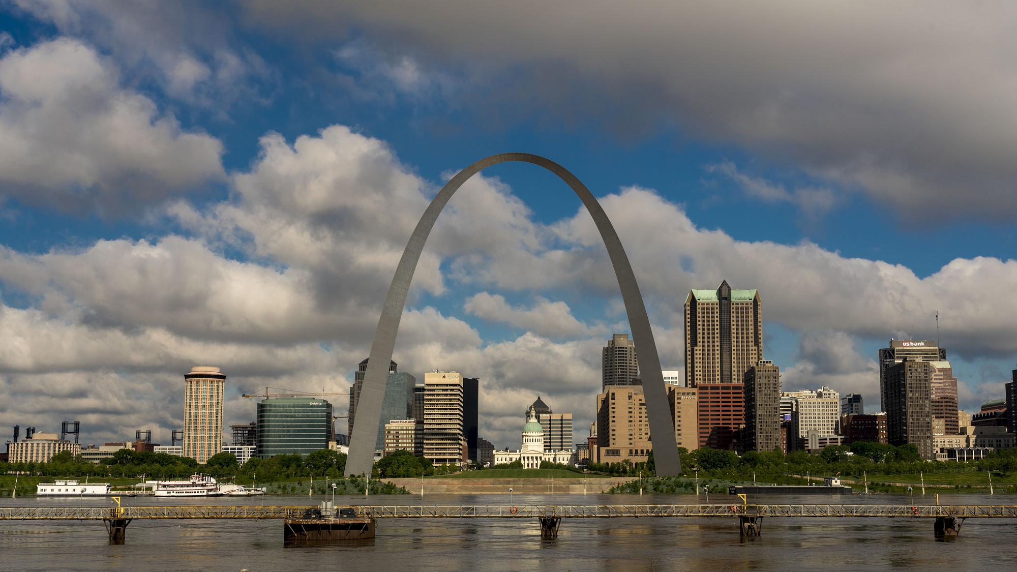 The Mississippi River is visible in the foreground of a photo, with the Gateway Arch in the center.  Buildings are in the background, and a blue sky with clouds is visible. 